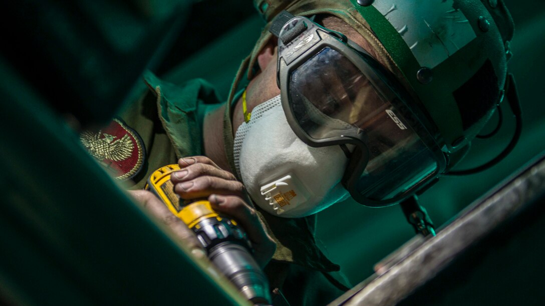 A Marine wearing protective goggles tilts his head and uses a tool to perform maintenance.