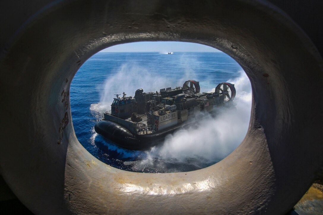 An air-cushioned landing craft, visible through a circular opening in a nearby ship, travels in blue waters.