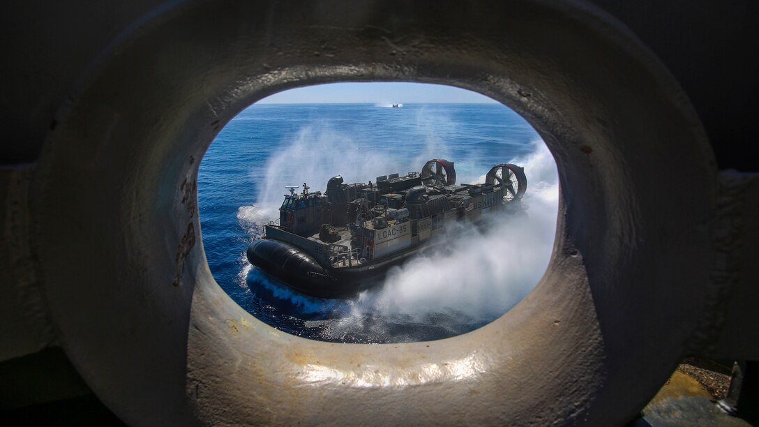 An air-cushioned landing craft, visible through a circular opening in a nearby ship, travels in blue waters.