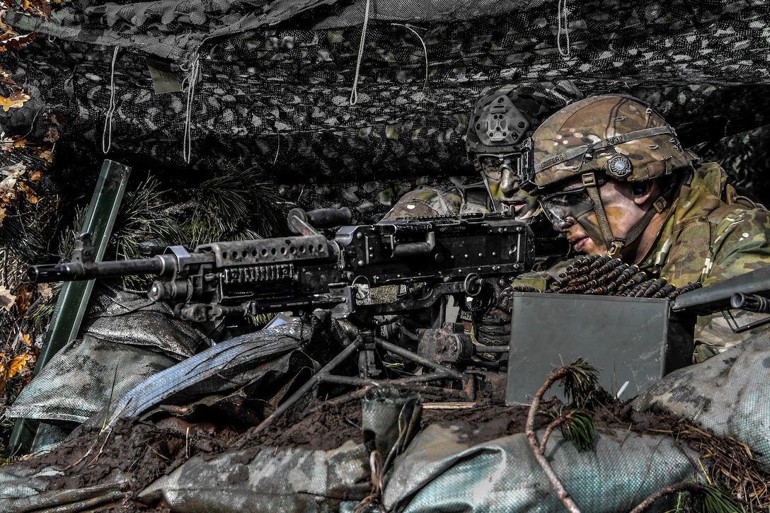A soldier looks through a gun's scope while crouched by another soldier against a backdrop of netting.
