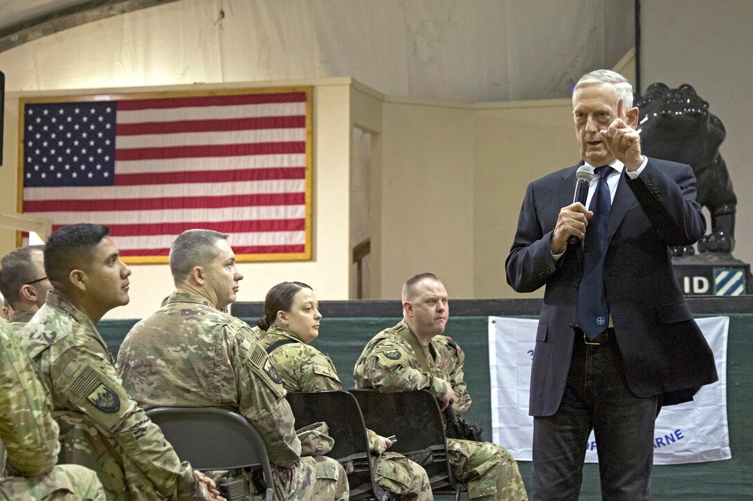Defense Secretary James N. Mattis stands and talks to seated troops in a room with a U.S. flag on the wall.