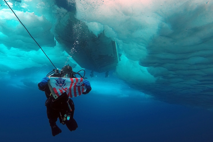 Coast Guard Petty Officer 2nd Class Adam Harris, a member of a joint Coast Guard-Navy dive team deployed on the Coast Guard Cutter Healy, holds a Coast Guard ensign during a cold water ice dive off a Healy small boat in the Arctic, July 29, 2017.