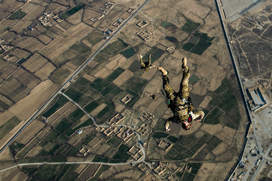 Three airmen free-fall with their limbs splayed before their parachutes have opened over brown and green terrain.