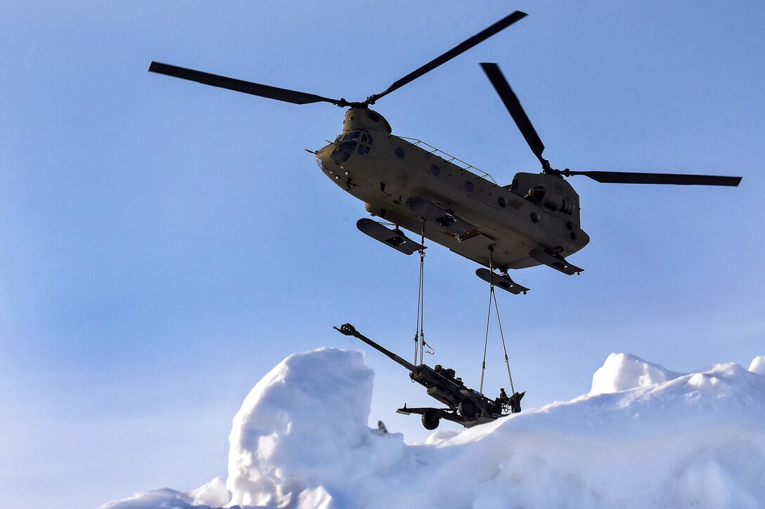 A helicopter delivers a howitzer to a snowy training area.