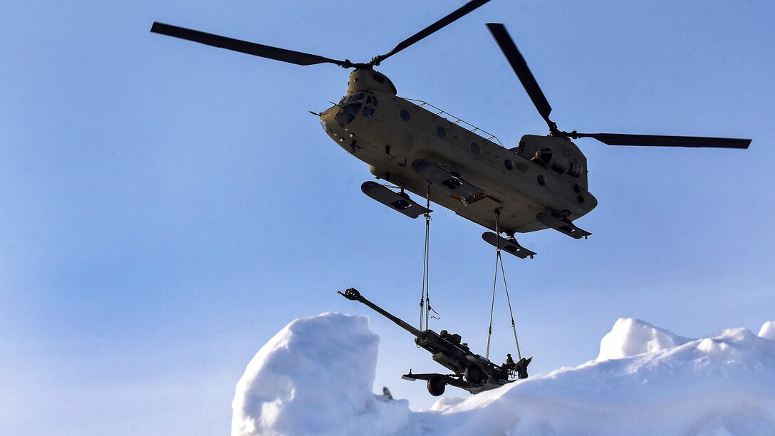 A helicopter delivers a howitzer to a snowy training area.