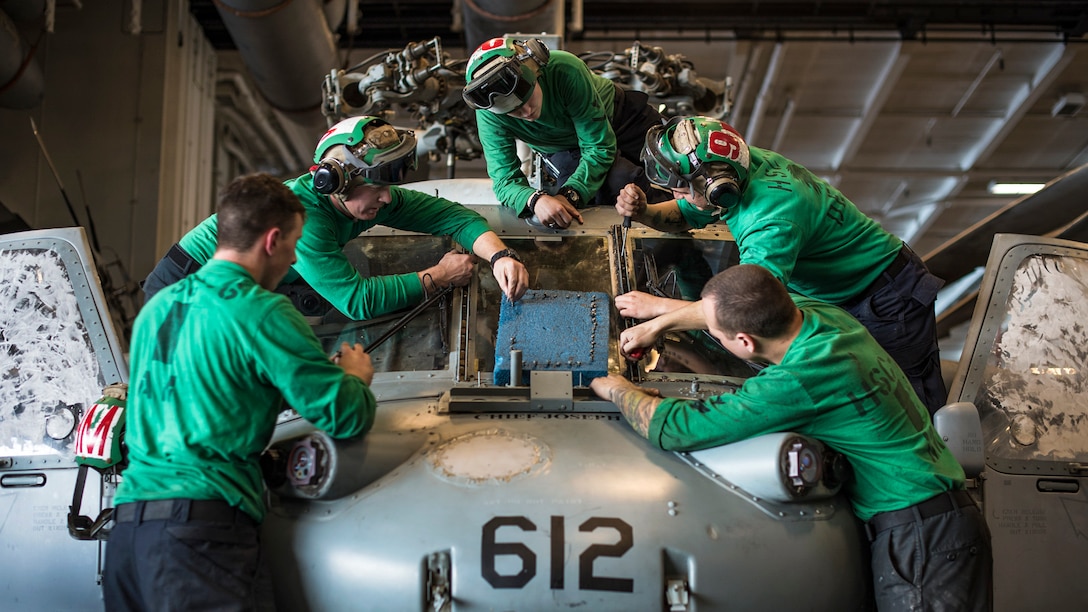 Sailors in green tops replace a windshielf on a helicopter on a ship.