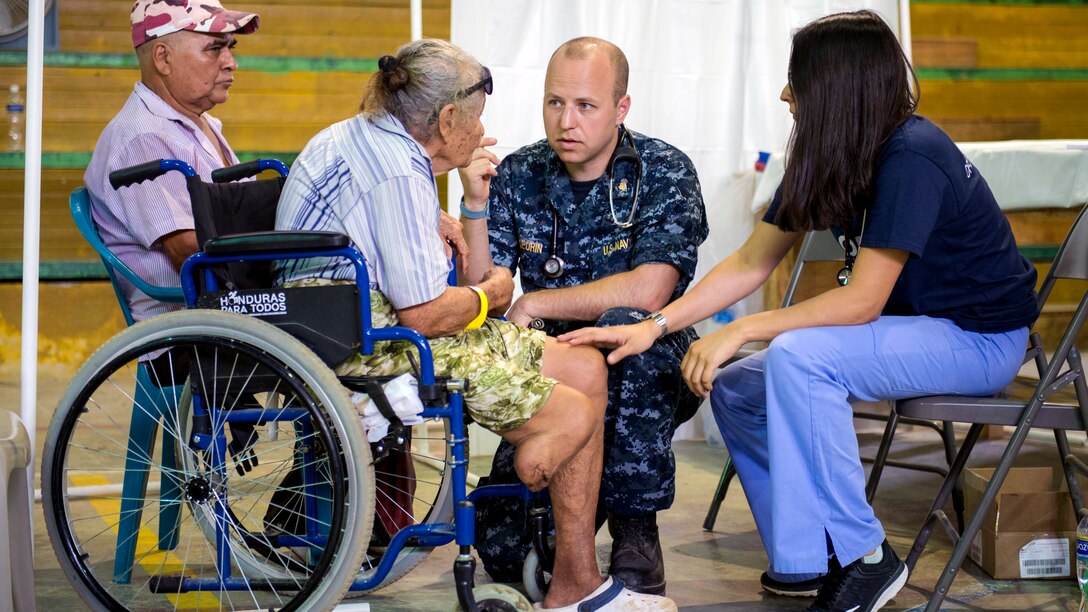 A sailor kneels while talking to an elderly woman in a wheelchair.