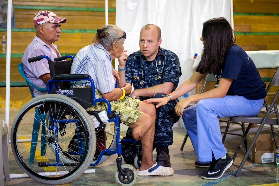 A sailor kneels while talking to an elderly woman in a wheelchair.