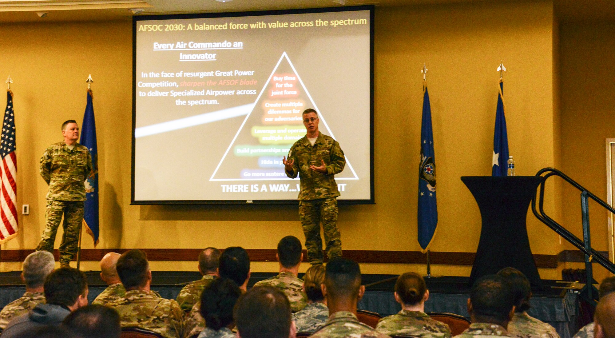 Two people speaking to a crowd of people with a screen and flags in the background.