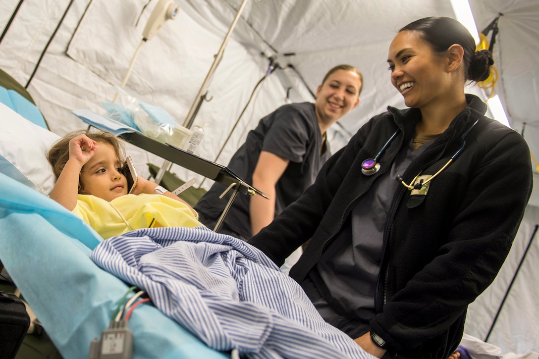 A sailor smiles at a Honduran girl listening to a cell phone in a hospital bed before surgery.
