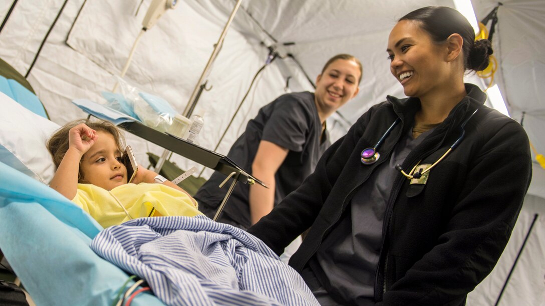 A sailor smiles at a Honduran girl listening to a cell phone in a hospital bed before surgery.