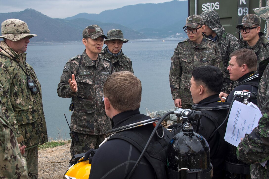 South Korean and U.S troops with diving equipment listen to a man talking.