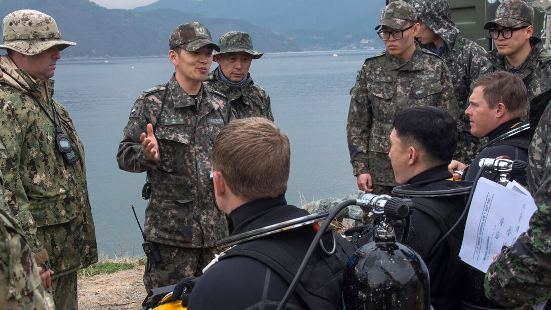 South Korean and U.S troops with diving equipment listen to a man talking.