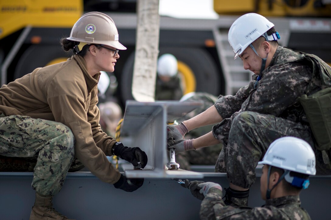 A U.S. sailor and and South Korean sailor sit across from each other placing an I-beam.