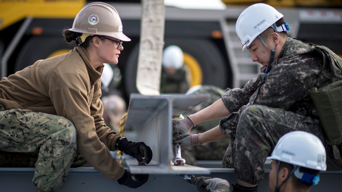 A U.S. sailor and and South Korean sailor sit across from each other placing an I-beam.