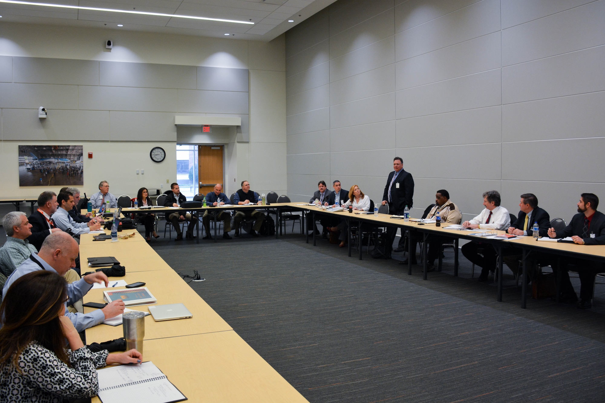 Approximately 20 people surround a u-shaped table. A man is standing and presenting to the group.