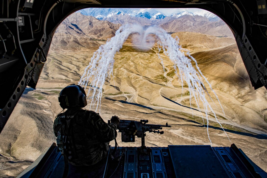 A soldier sits facing out from an open helicopter as two curving lines of smoke trails waft over brown hilly terrain.