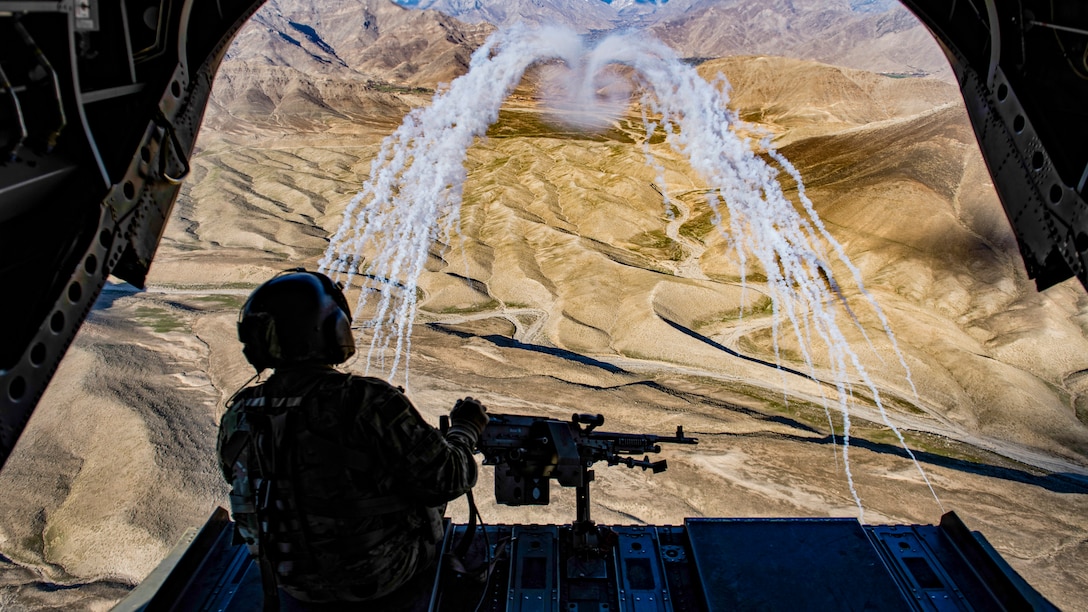 A soldier sits facing out from an open helicopter as two curving lines of smoke trails waft over brown hilly terrain.