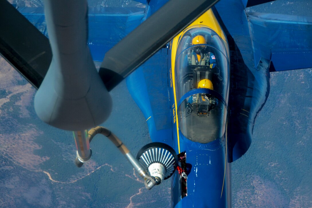 A fuel hose from one aircraft extends into a blue jet flying beneath it.