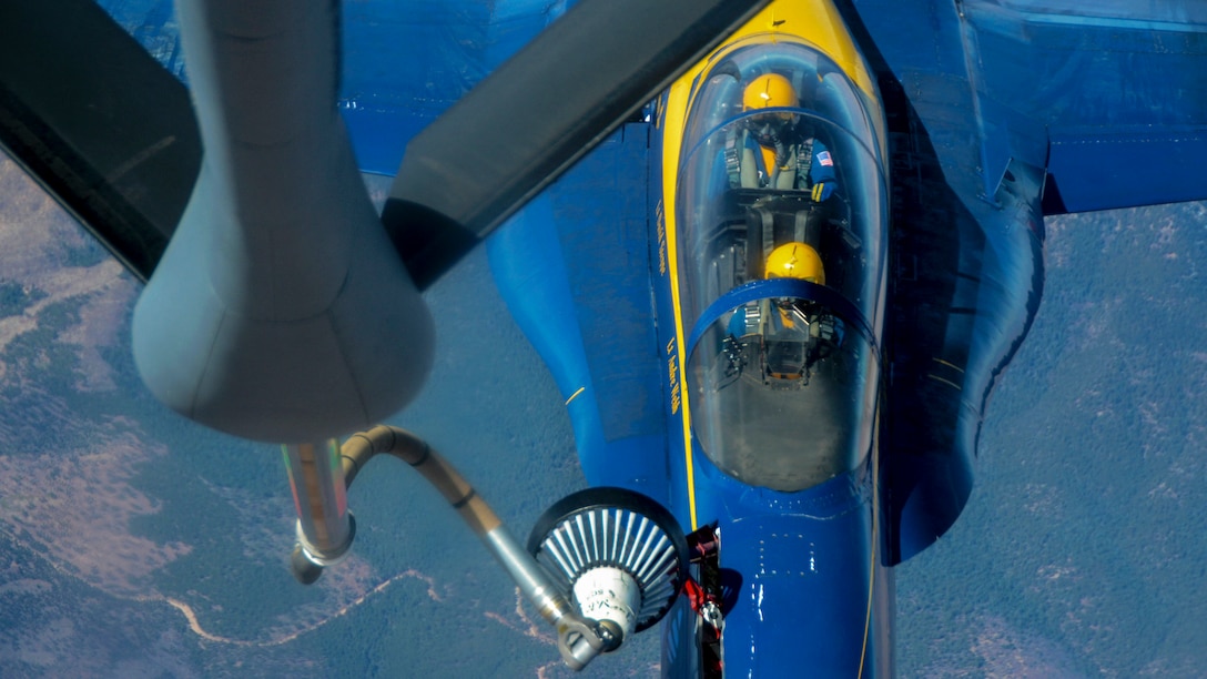 A fuel hose from one aircraft extends into a blue jet flying beneath it.