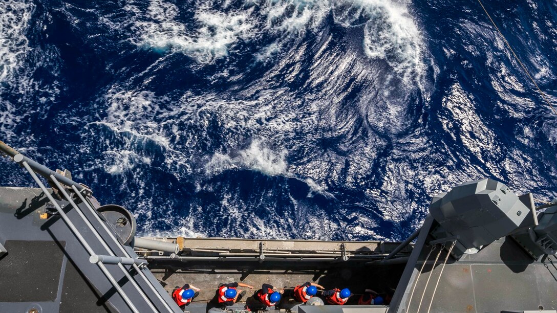 Sailors, shown from above, pull a rope on a ship traveling in roiling blue water.