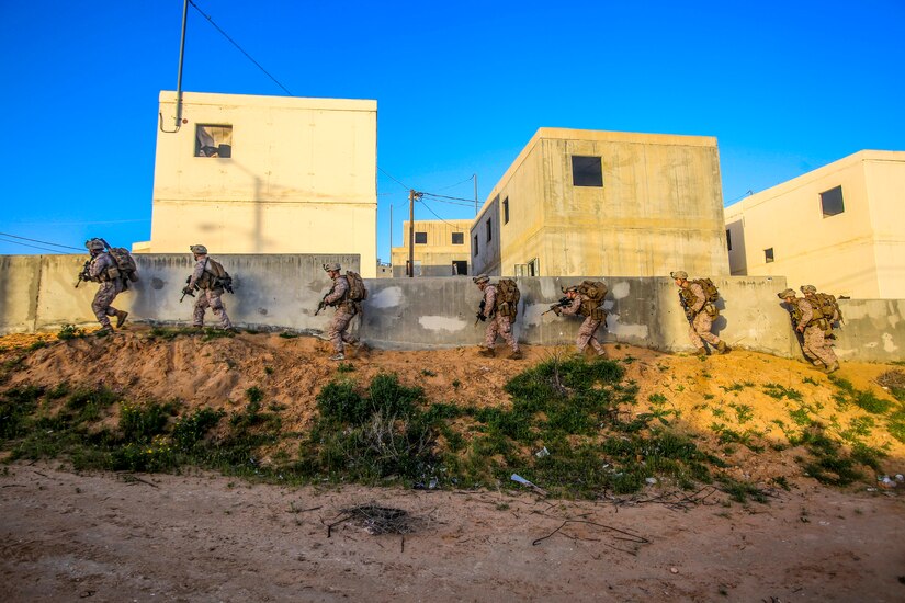Marines crouch and walk along a wall carrying weapons.