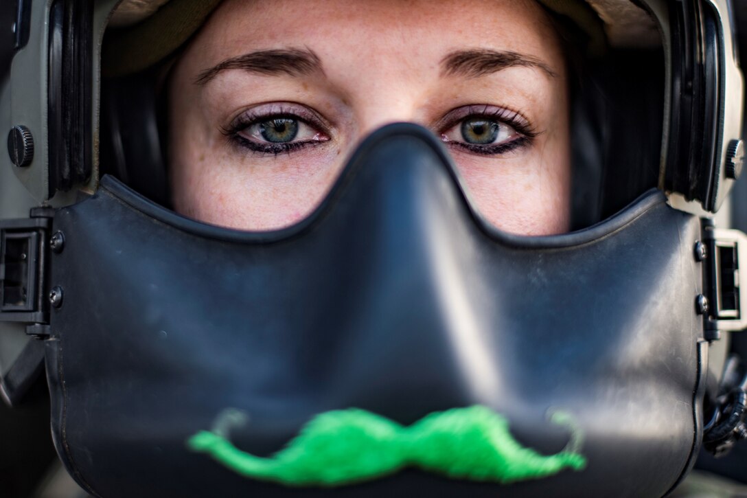 A closeup of a female airman with a green mustache stuck to her flying helmet.