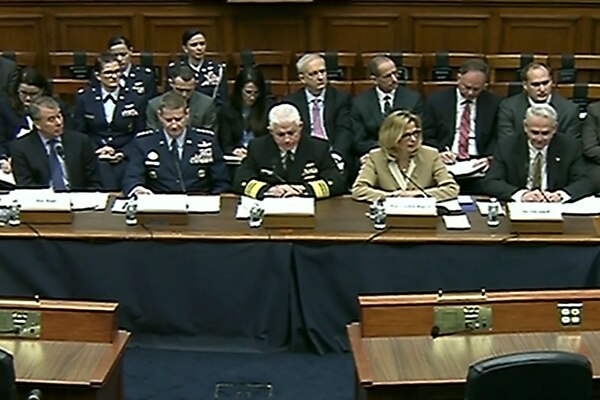 A row of civilians and military officers sit behind a desk.