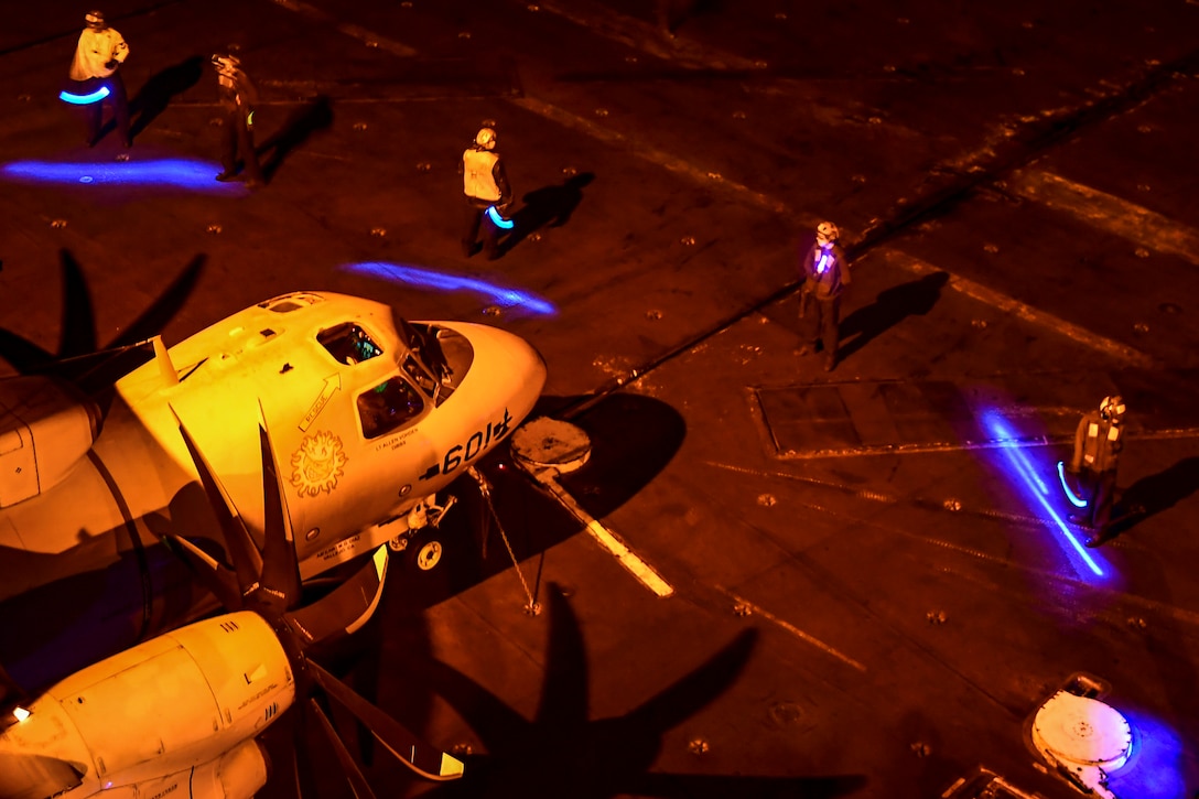 Sailors form a safety boundary around a helicopter on a ship's flight deck at night.