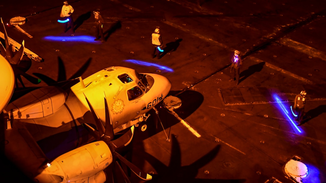 Sailors form a safety boundary around a helicopter on a ship's flight deck at night.