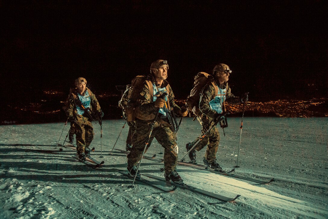 Three Marines cross-country ski in the dark on a snowy mountain summit.