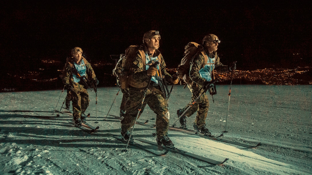 Three Marines cross-country ski in the dark on a snowy mountain summit.