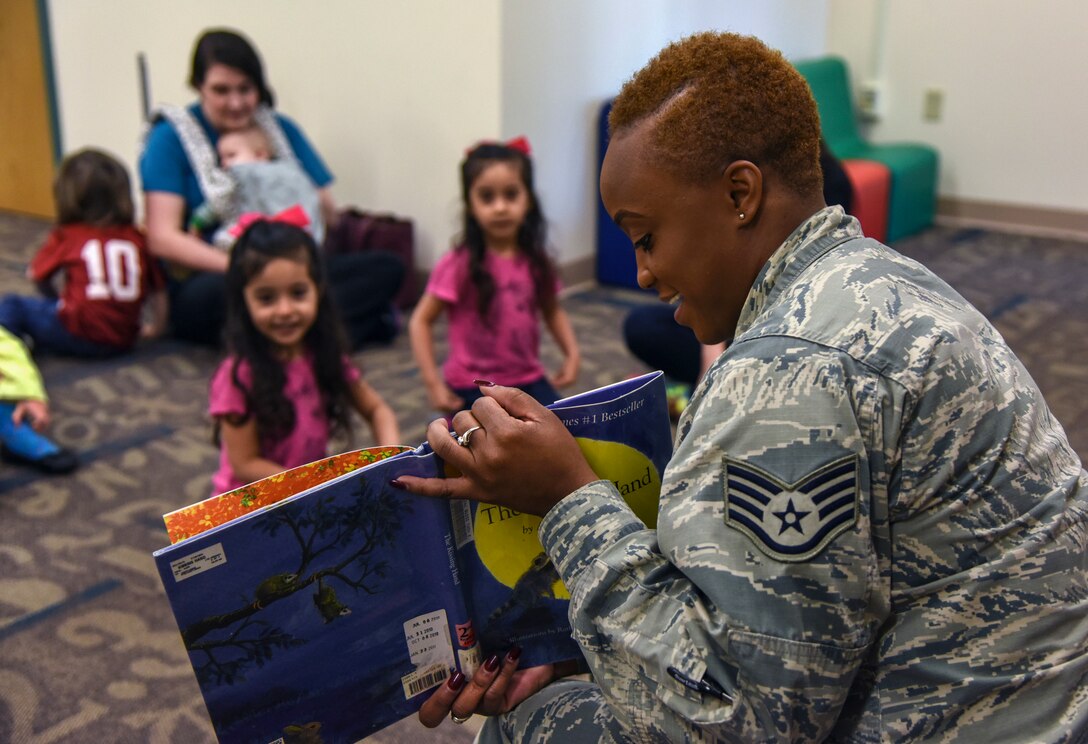 An airman smiles while reading a picture book to children, who smile while looking at the book.