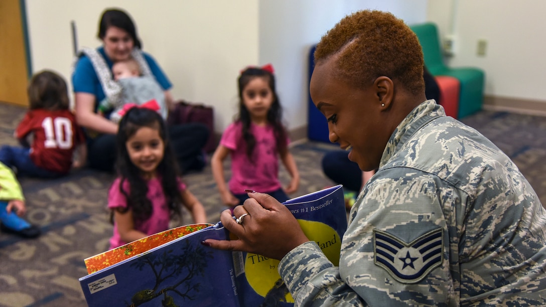 An airman smiles while reading a picture book to children, who smile while looking at the book.