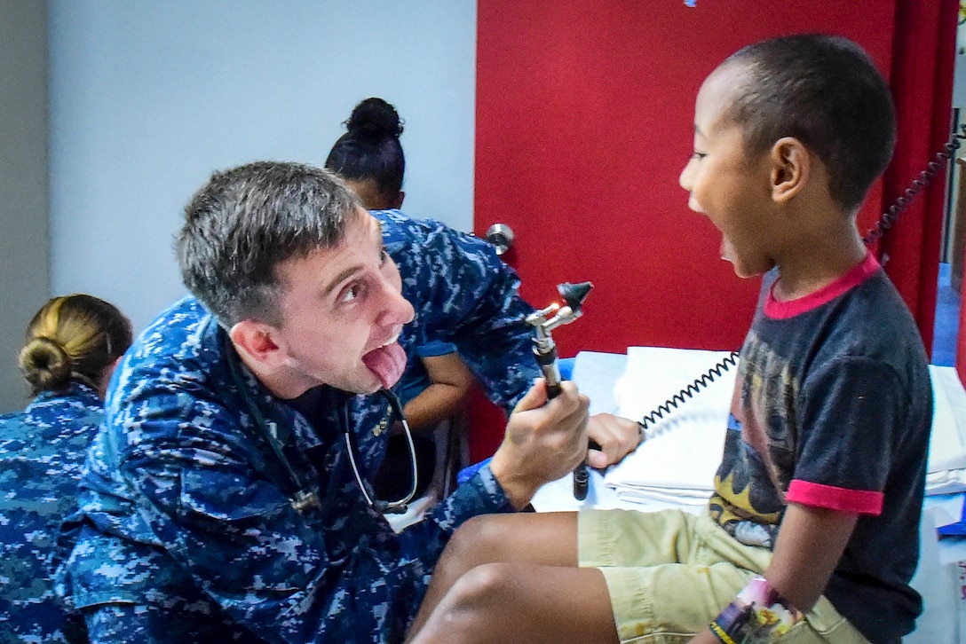A Navy doctor holding an otoscope sticks his tongue out at a boy, who opens his mouth wide for the doctor to examine his throat.