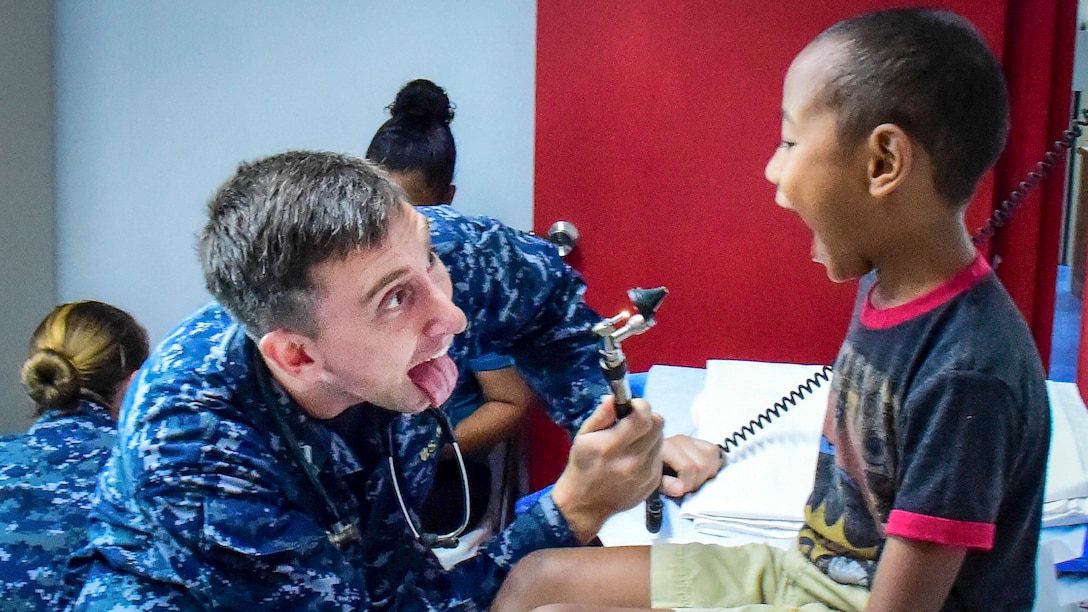 A Navy doctor holding an otoscope sticks his tongue out at a boy, who opens his mouth wide for the doctor to examine his throat.