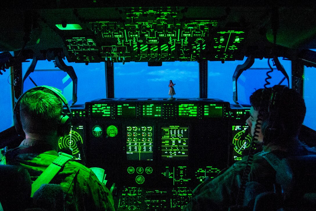 Airmen sit in a cockpit illuminated in green light, with turquoise sky showing through the windows.