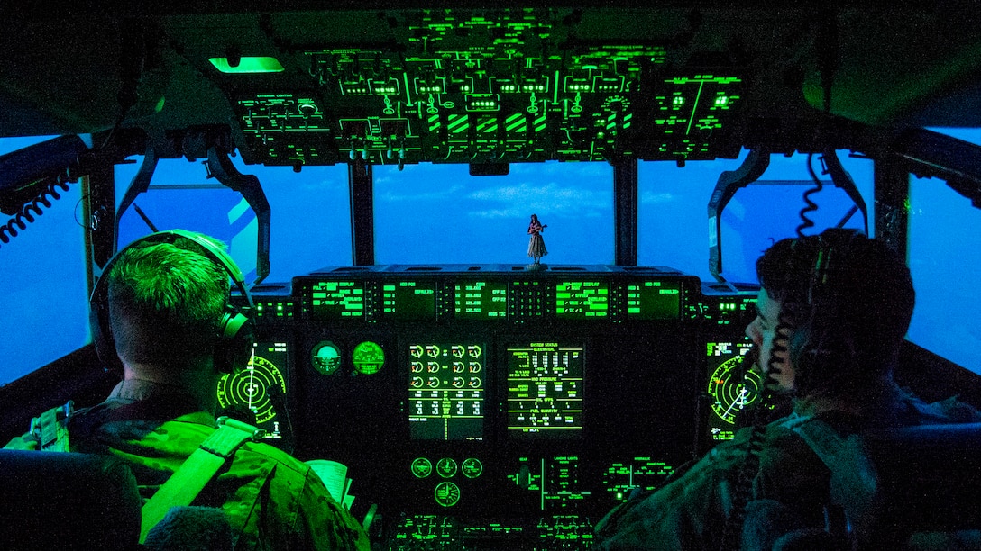 Airmen sit in a cockpit illuminated in green light, with turquoise sky showing through the windows.