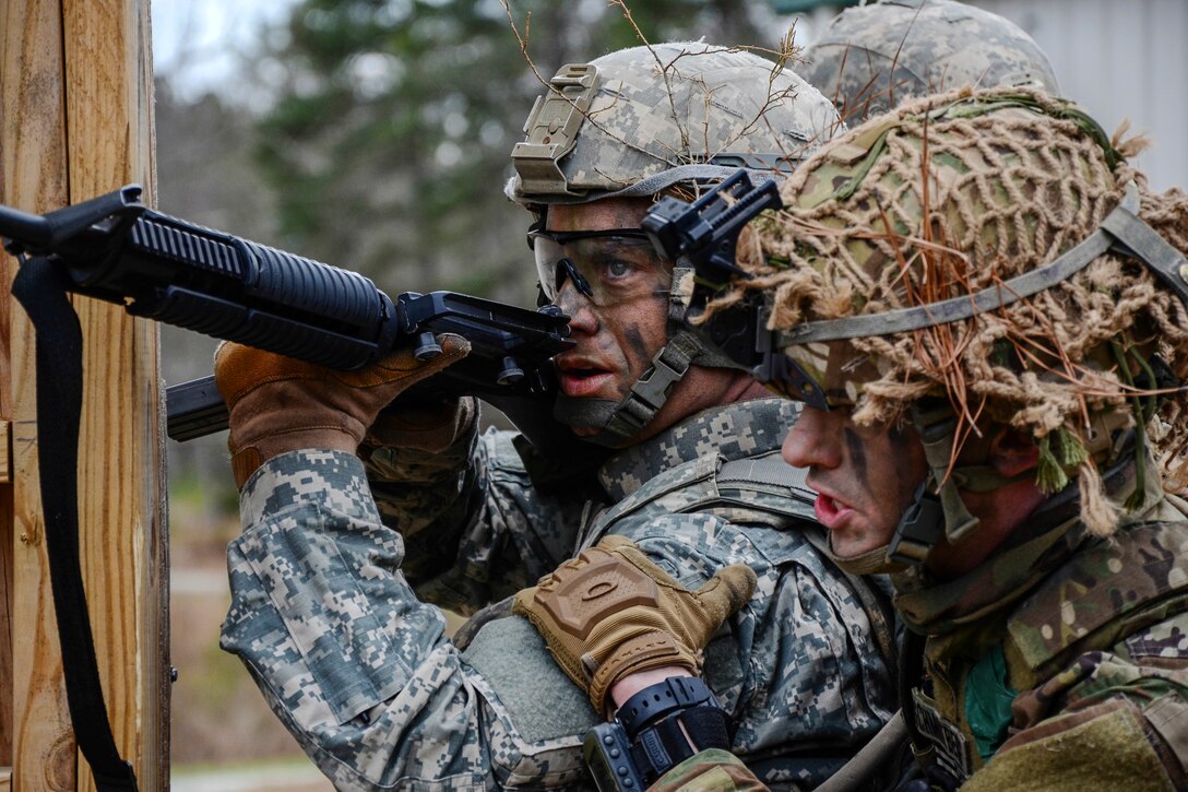 A soldier points a weapon as another stands right behind him, holds his arm and speaks to him.