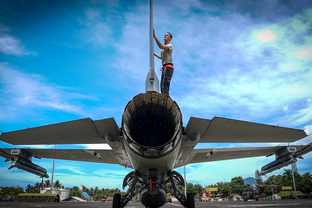 An airman stands atop a fighter jet and examines the tail.