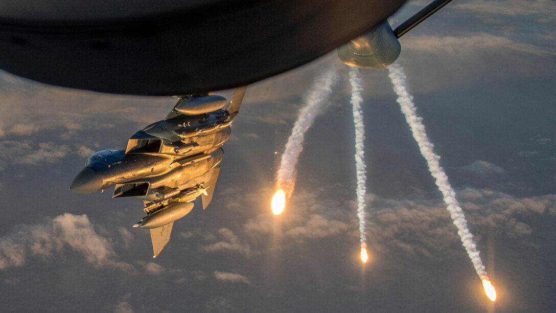 Three flares fire in the sky adjacent to a fighter jet flying below a tanker aircraft.