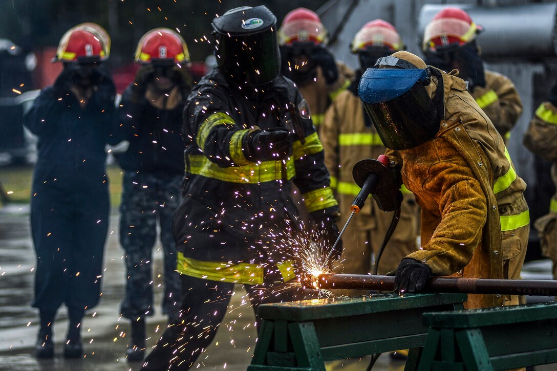 Sparks fly as a sailor in a protective mask uses a tool to cut metal, as an instructor gives directions.