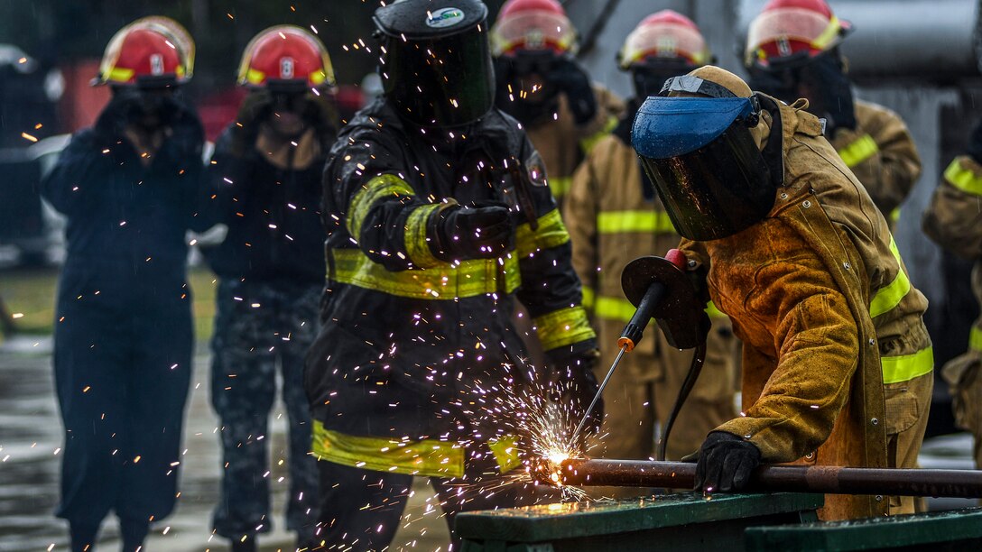 Sparks fly as a sailor in a protective mask uses a tool to cut metal, as an instructor gives directions.