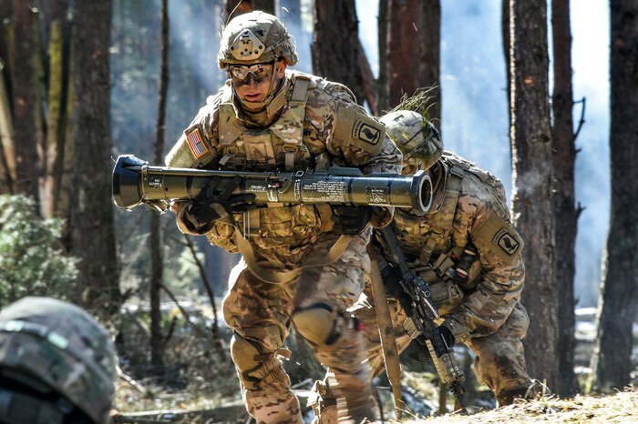 A soldier carries a grenade launcher while hurrying through a wooded area.