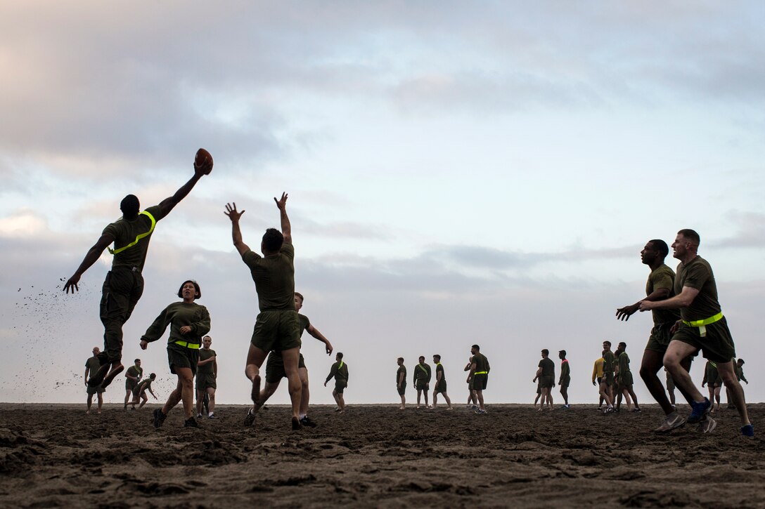 A Marine leaps and reaches his arm out to catch a football while on a beach with other players.