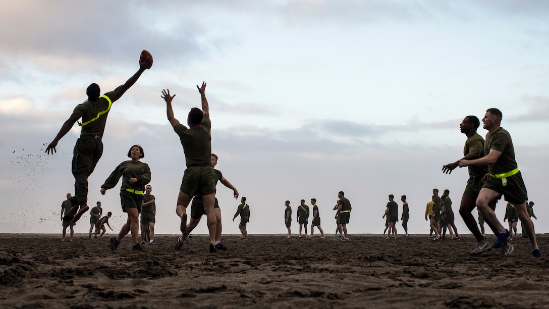 A Marine leaps and reaches his arm out to catch a football while on a beach with other players.