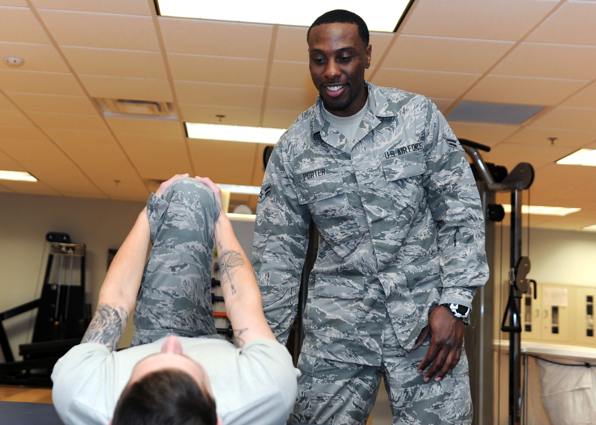 A male standing in a room with exercise equipment helps another male stretch.