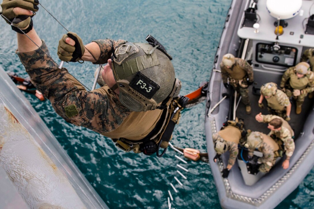 A Marine climbs a rope ladder over water, as troops stand by in a rubber boat below.