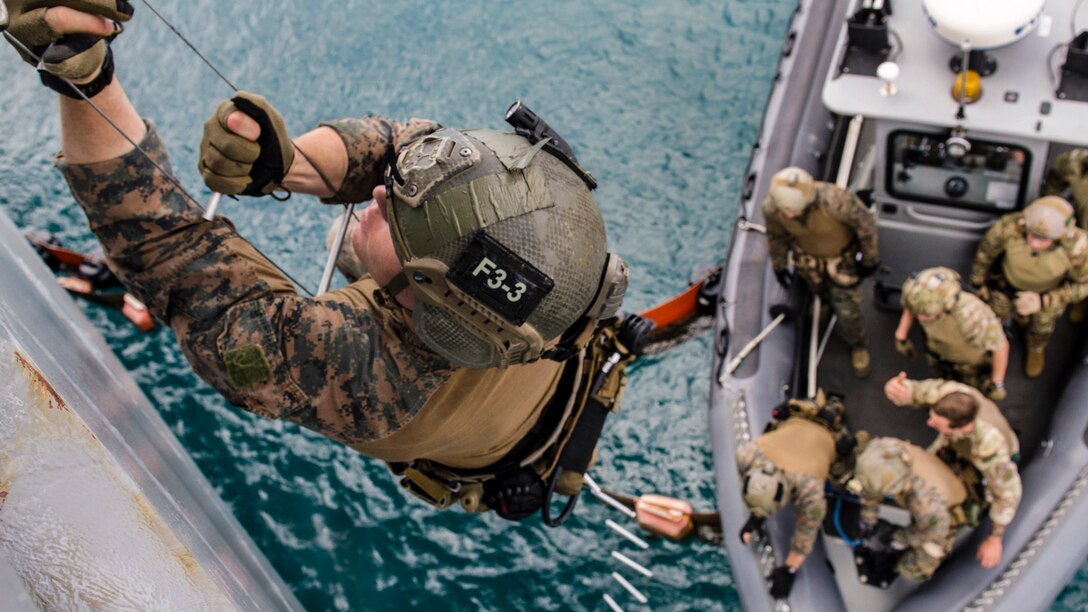 A Marine climbs a rope ladder over water, as troops stand by in a rubber boat below.