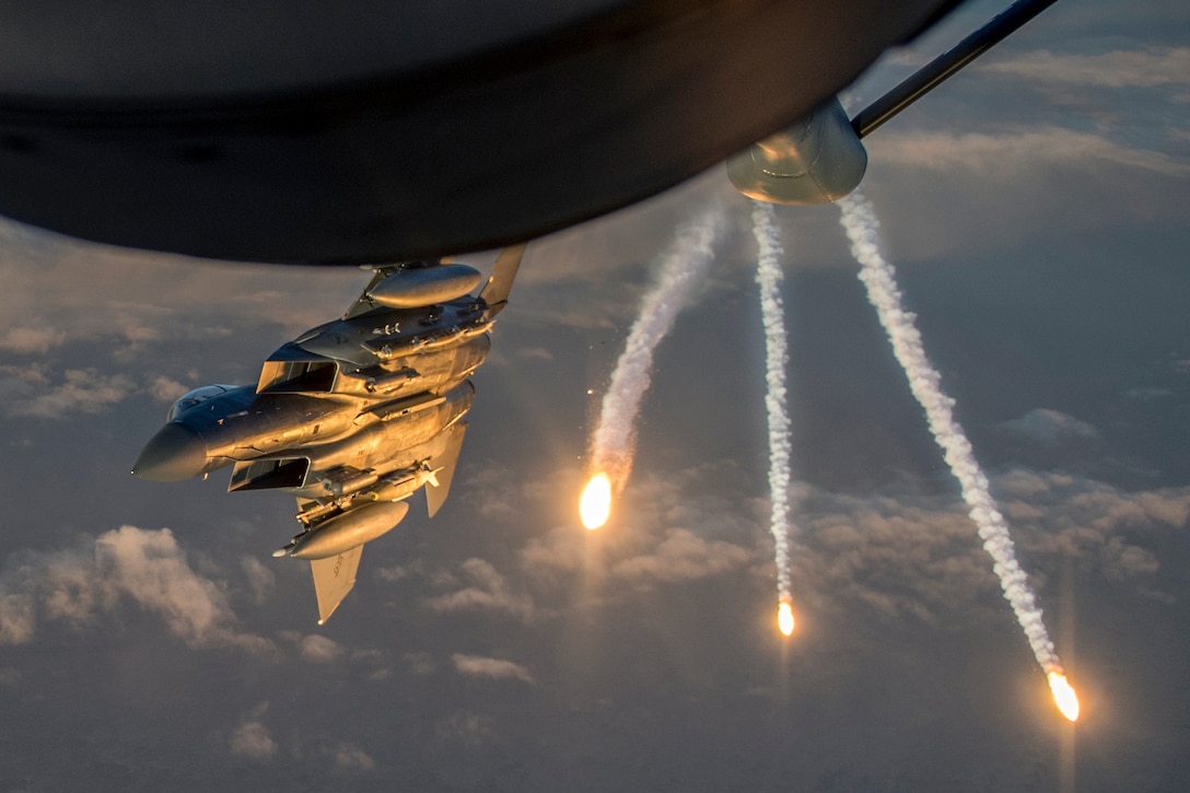 Three flares fire in the sky adjacent to a fighter jet flying below a tanker aircraft.